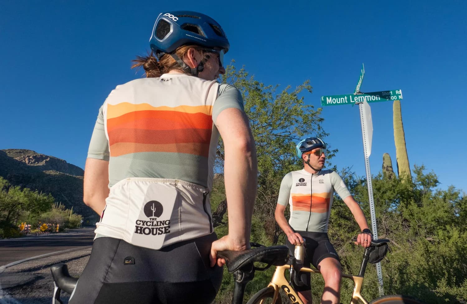 Two cyclists wearing The Cycling House desert-tone kits beside the Mount Lemmon road sign